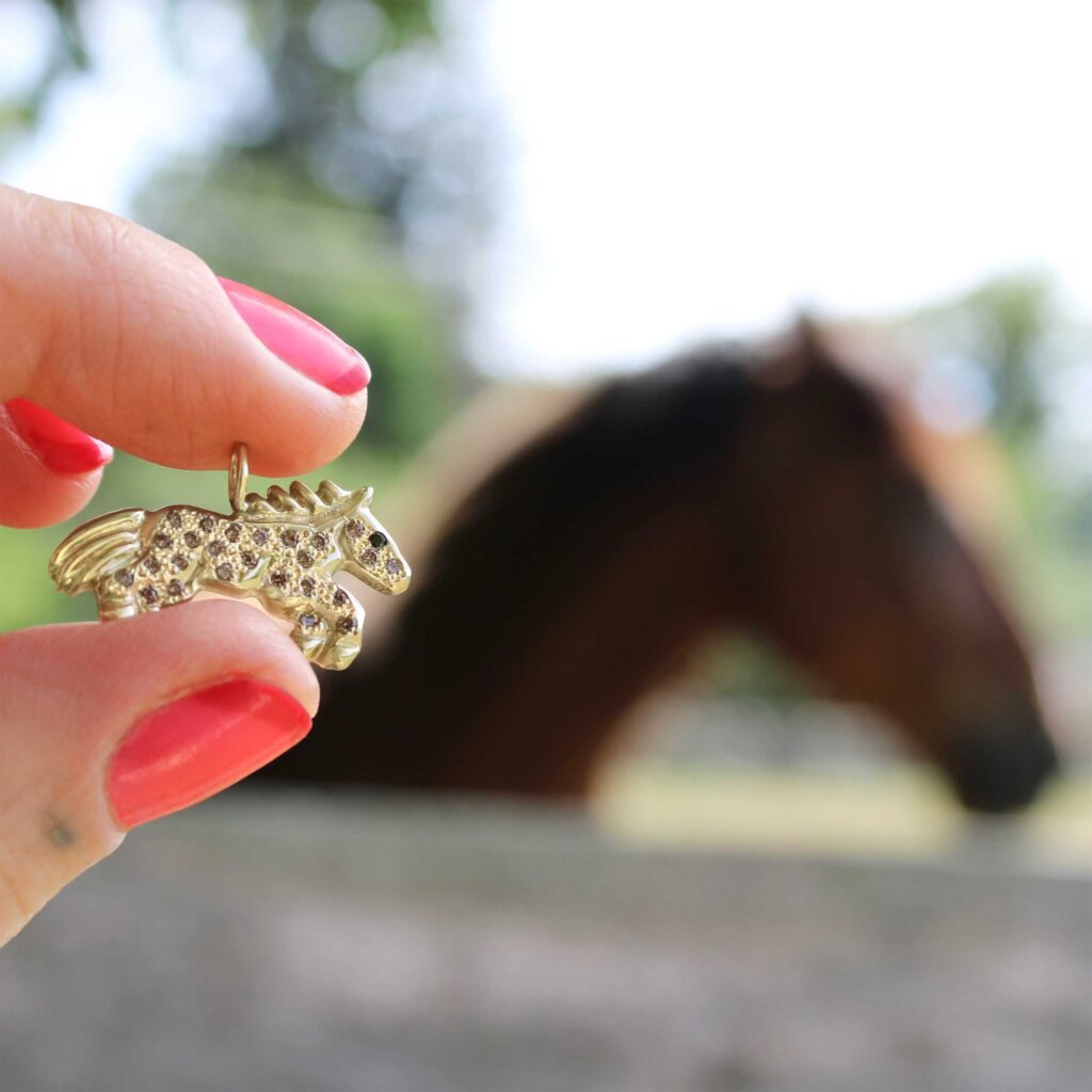 holding up a gold horse charm in the foreground with a horse in the background